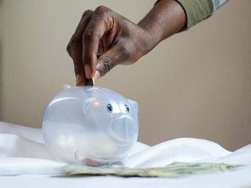 Close-up of a person's hand placing coins into a transparent piggy bank to save money.